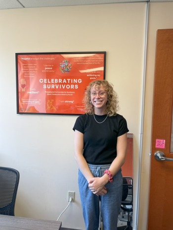 Emma Gross '25 stands in front of a poster at her internship that reads "Celebrating Survivors"