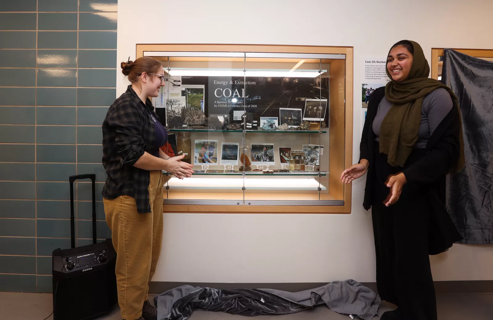 Wall display case containing photos and geological specimens presented on either side by two students. 