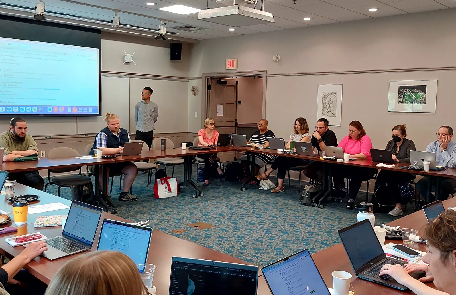 A room full of people with laptops listening to a speaker