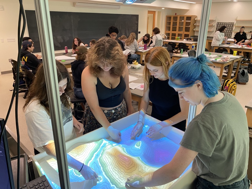 Four students stand around the augmented reality sandbox, moving the sand to create new terrain on top of it