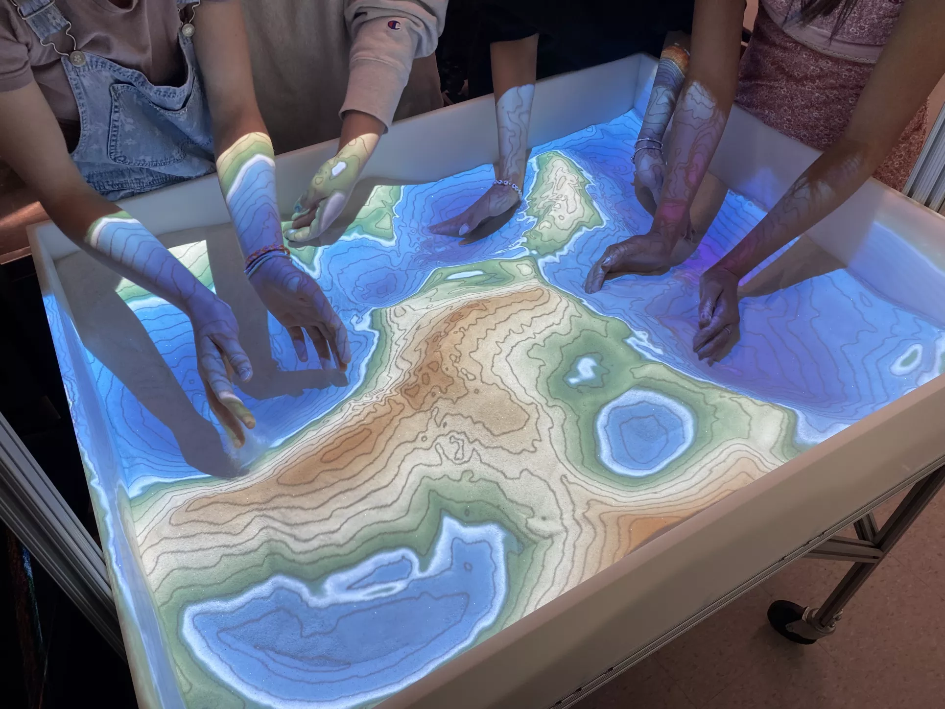 Several students put their hands into the sandbox. The digital map projected on the sand changes as the students move the sand around.