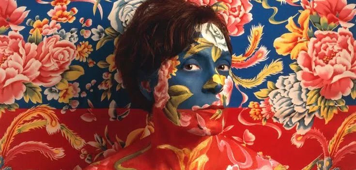 Self Portrait of Artist wearing blue and red floral make-up as camouflage with wallpaper behind her