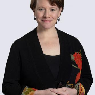 A woman with short, brown hair smiles at the camera. She is wearing a black shirt and a black blazer with embroidered cuffs.