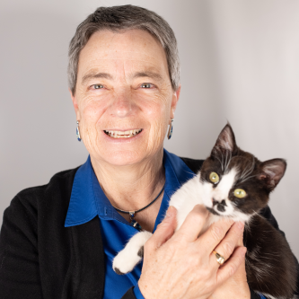 A woman with short dark grey hair smiles at the camera. She is wearing a cobalt blue button down shirt and a black cardigan. She is holding a black and white cat, who is looking off to the side.