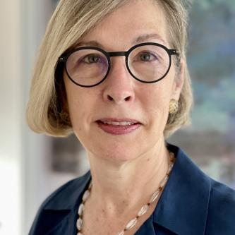 A woman with jaw length blond hair looks at the camera. She is wearing round glasses, a navy blazer, and a pink shirt.