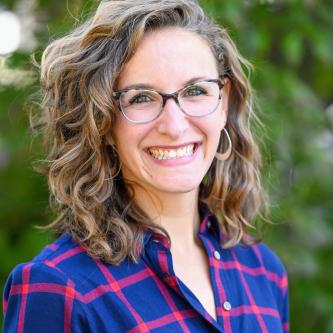 A woman with blond wavy hair smiles at the camera. She is standing in front of greenery. She is wearing a blue and red plaid shirt, glasses, and silver hoop earrings.