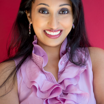 A woman with long black hair smiles at the camera, in front of a hot pink background. She is wearing a pink ruffled shirt and dangly earrings.