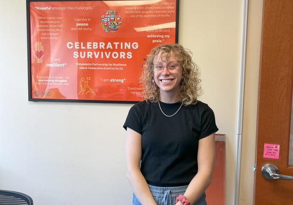 Emma Gross '25 stands in front of a poster at her internship that reads "Celebrating Survivors"