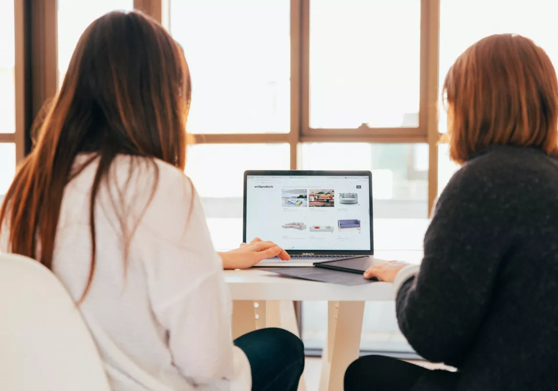 Two persons look at content displayed in a laptop. Their back is to the camera.