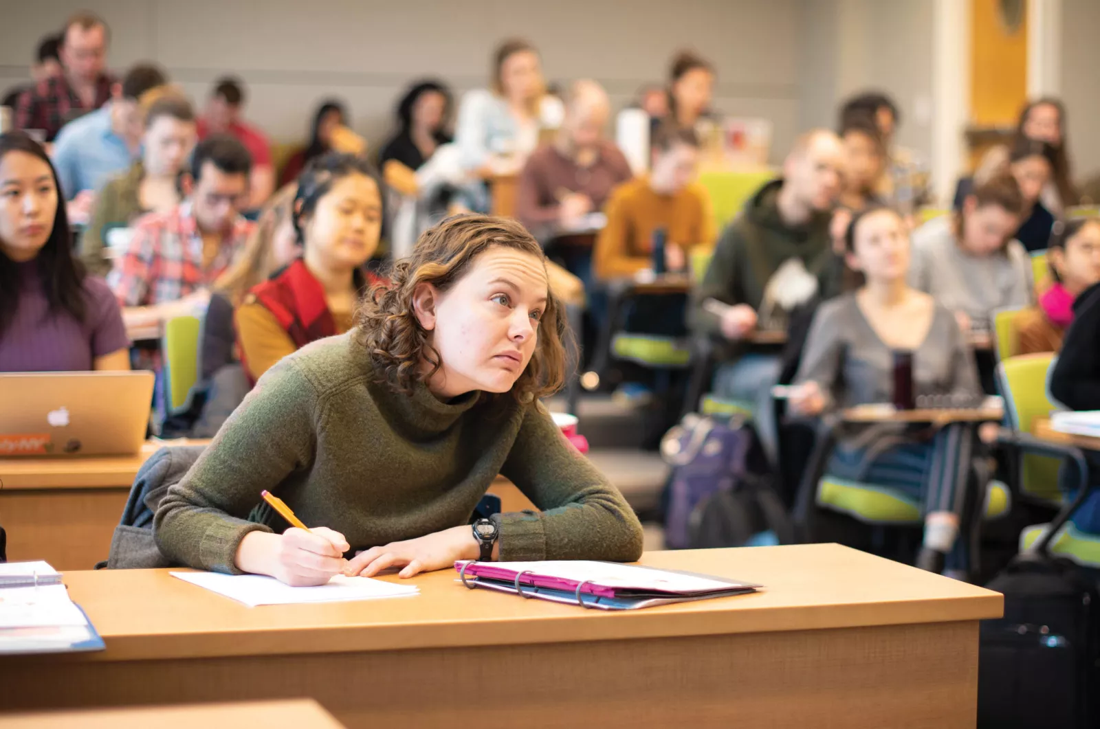 A class of students with one student in focus. That student is taking notes, looking toward the front of the room.