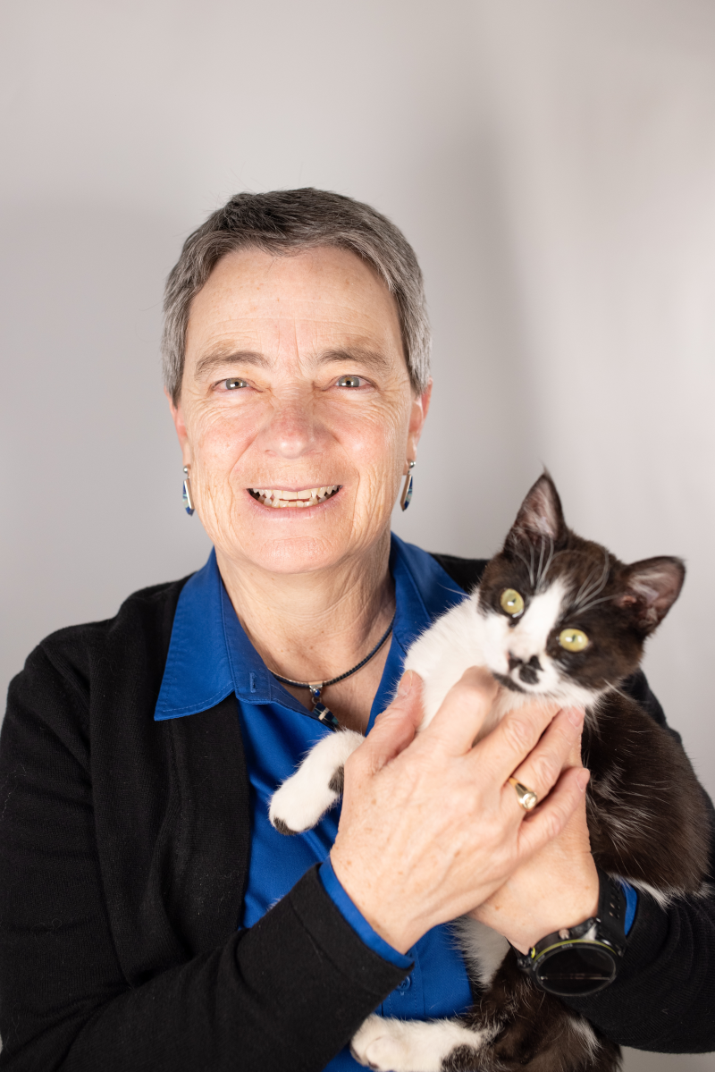 A woman with short dark grey hair smiles at the camera. She is wearing a cobalt blue button down shirt and a black cardigan. She is holding a black and white cat, who is looking off to the side.