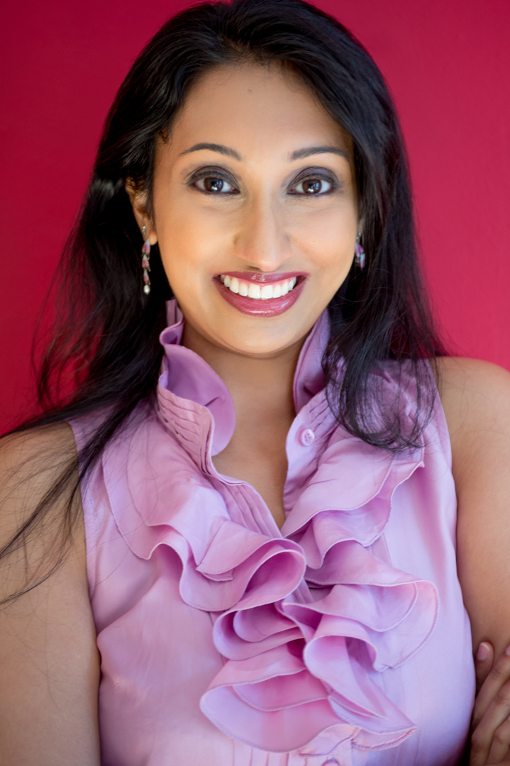 A woman with long black hair smiles at the camera, in front of a hot pink background. She is wearing a pink ruffled shirt and dangly earrings.